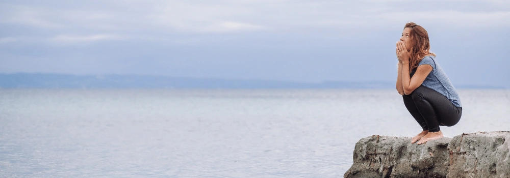 Kvinna som sitter på huk på liten klippa vid havet och tittar uppåt